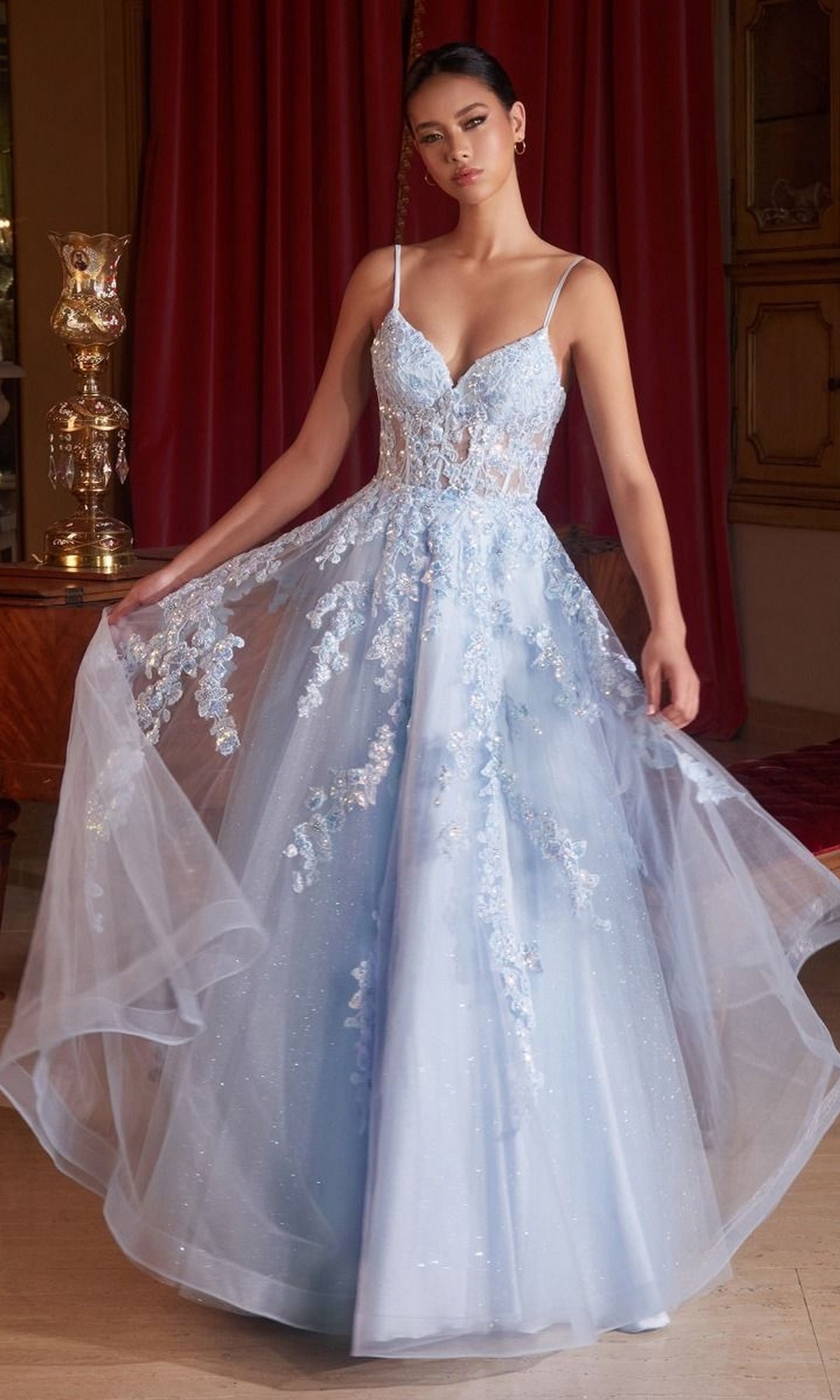 A woman models the Long Prom Ball Gown with Flowers CM347, featuring light blue fabric, floral embellishments, thin straps, lace appliqué, and a sheer tulle skirt. She stands indoors before red curtains in this elegant formal evening look.